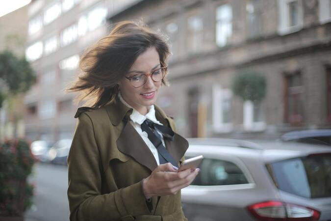 Eine jüngere Frau hält ein Smartphone in der Hand, lächelt leicht, ihr Blick ist auf das Smartphone gerichtet, der Wind luftet leicht im Haar, sie trägt einen Trenchcoat und im Hintergrund ist ein parkiertes Auto zu sehen. Sie läuft eine Häuserfassade entlang.